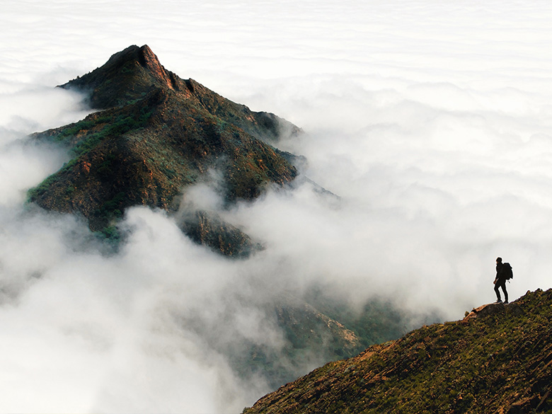 Ein Bergsteiger blickt auf einen Berggipfel, der sich über einer dicken Wolkendecke erhebt.
